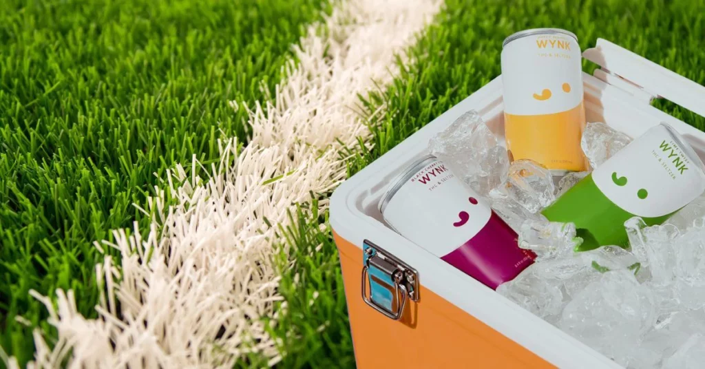 hree colorful cans of Wynk beverages chilled in an ice-filled cooler placed on green grass near a white field line